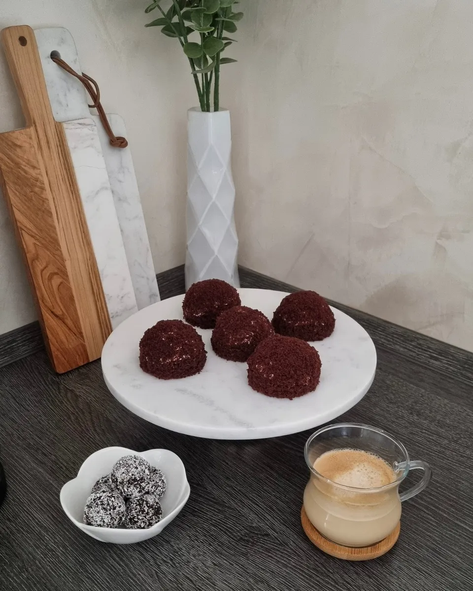 Cinq dômes de gâteau au cacao roulés dans les miettes, sur un présentoir de marbre, avec un bol de boules à la noix de coco et un café.