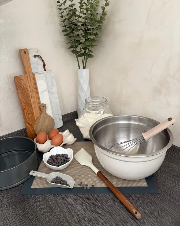 Un coin de plan de travail dressé avant de pâtisser : planches en bois et en marbre appuyées au mur, bocal de farine, trois œufs, pépites de chocolat, cul-de-poule et fouet, moule à charnière et rouleau.